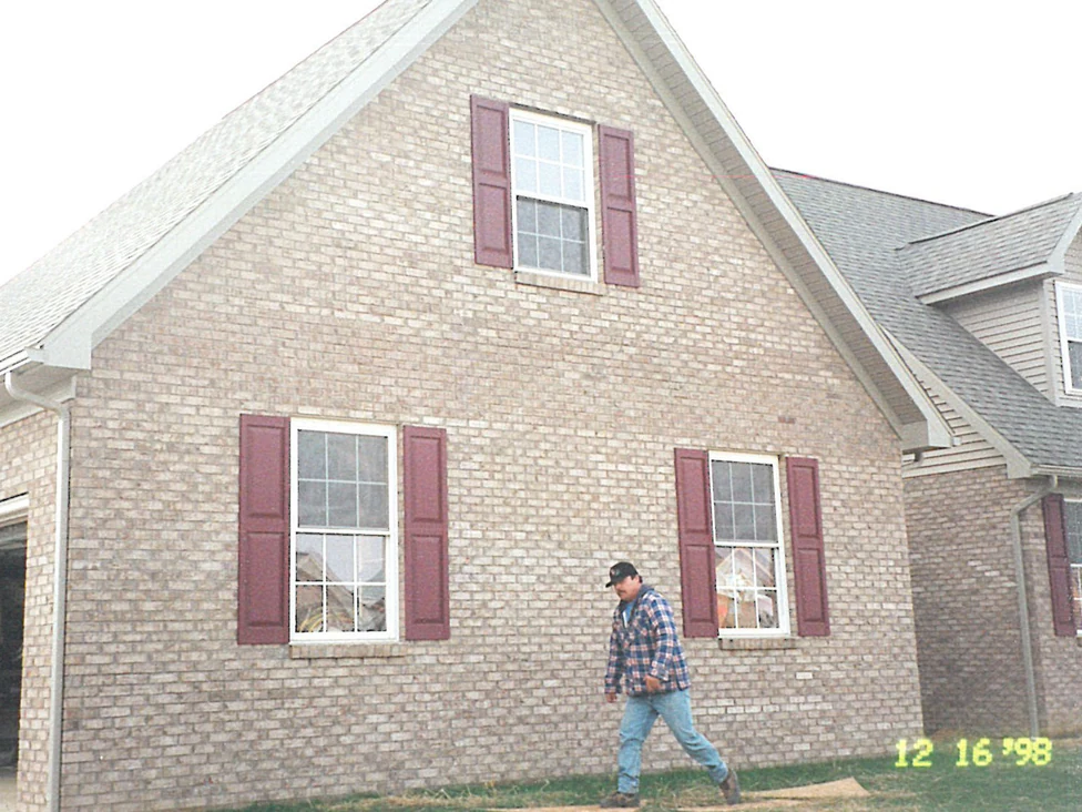 The garage end of the house in 1998 with a man walking past it, the brick a pale cream and tan under maroon shutters, the lot bare