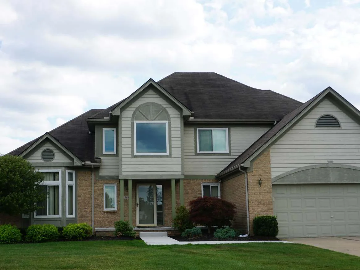 The front of the two-story house in 2019, tan brick on the ground floor under gray green siding and a dark roof, the brick one even range from the bay window past the entrance to the garage