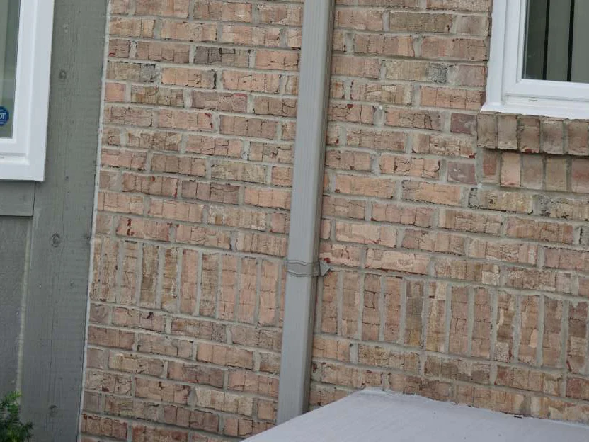 The brick beside a window and a downspout in 2019, tan and pink units under a rowlock sill with gray green siding above