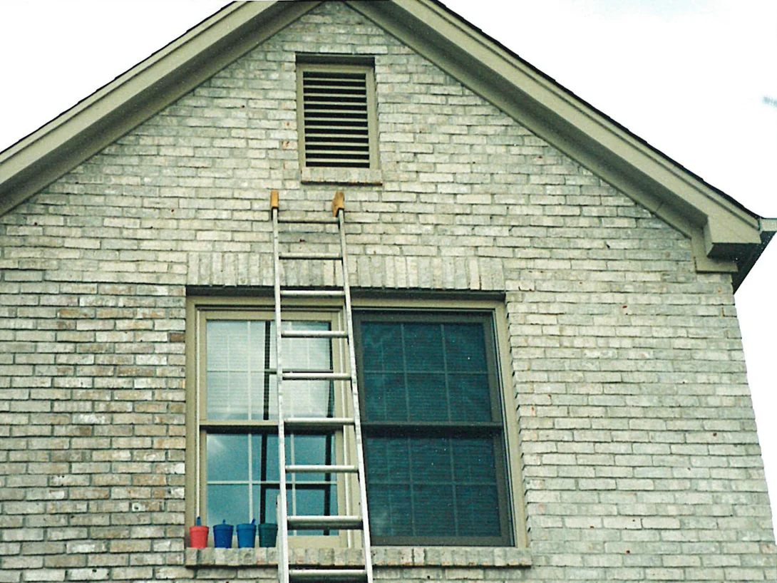 An extension ladder standing against the brick gable end of a house in 1998, an attic vent in the peak above a wide window, the brick a pale grey and cream range