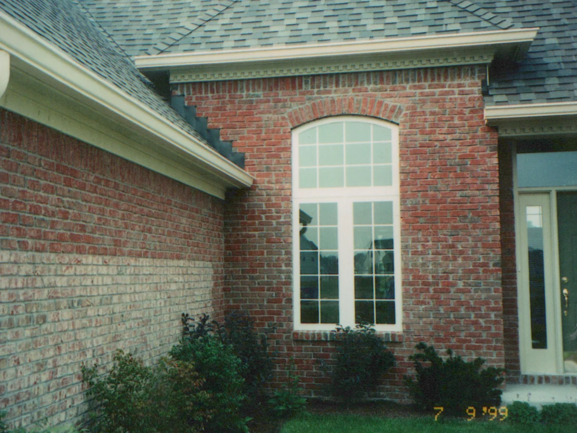 The side wall of the house in 1999, red brick above and a band of paler, pinker brick through the lower courses, beside an arched window with white trim and a dentil cornice