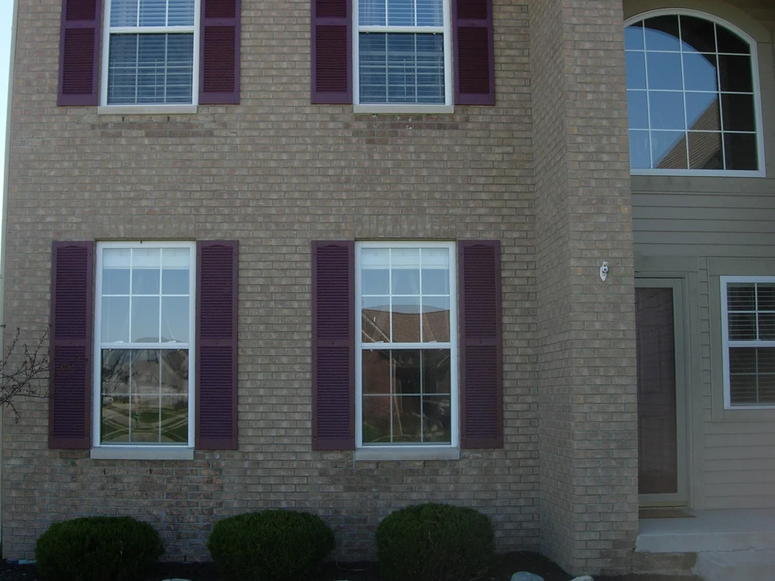 The corner of a two-story buff brick house in 2007, a run of darker grey brown brick sitting under both lower windows above the shrub bed