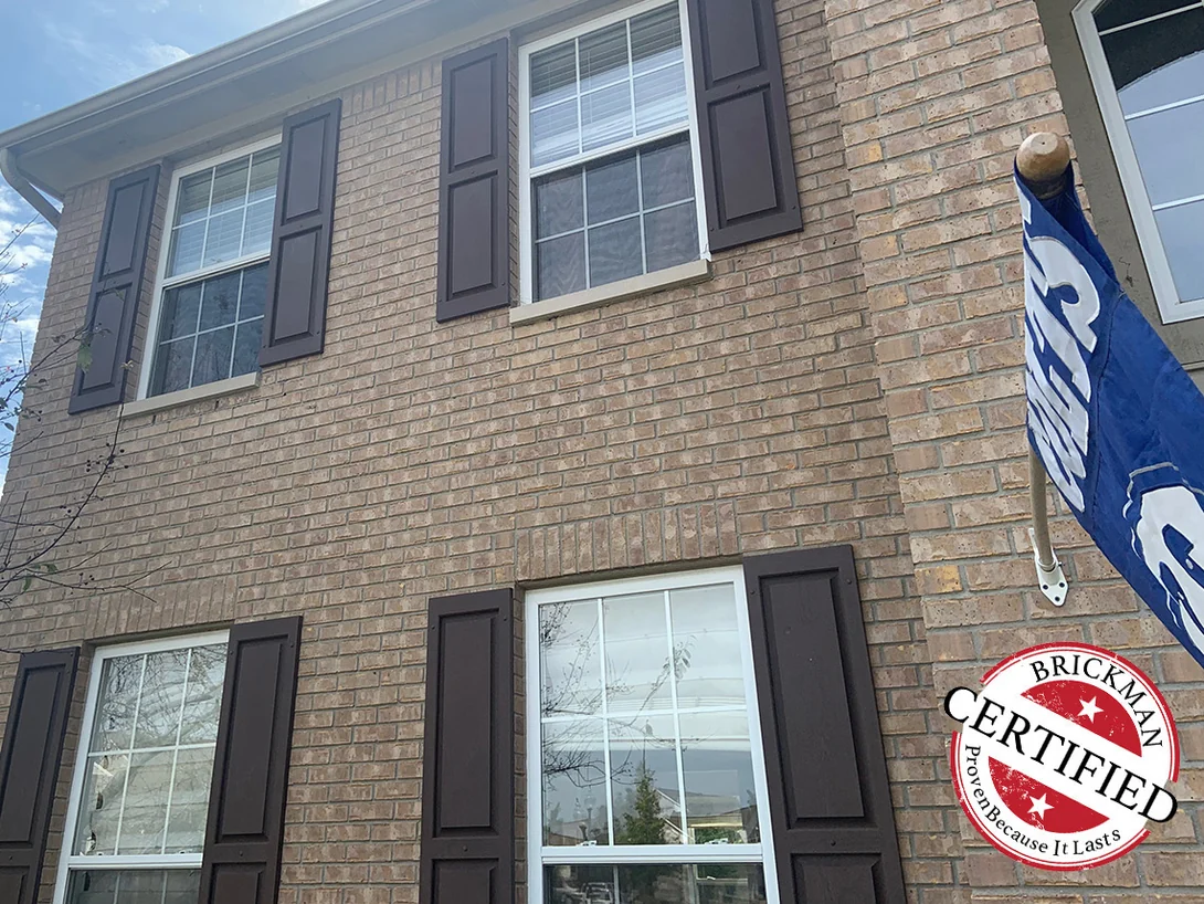 The front of the house in 2019 from the lawn, two floors of brick running past shuttered windows, with a certification stamp in the lower corner
