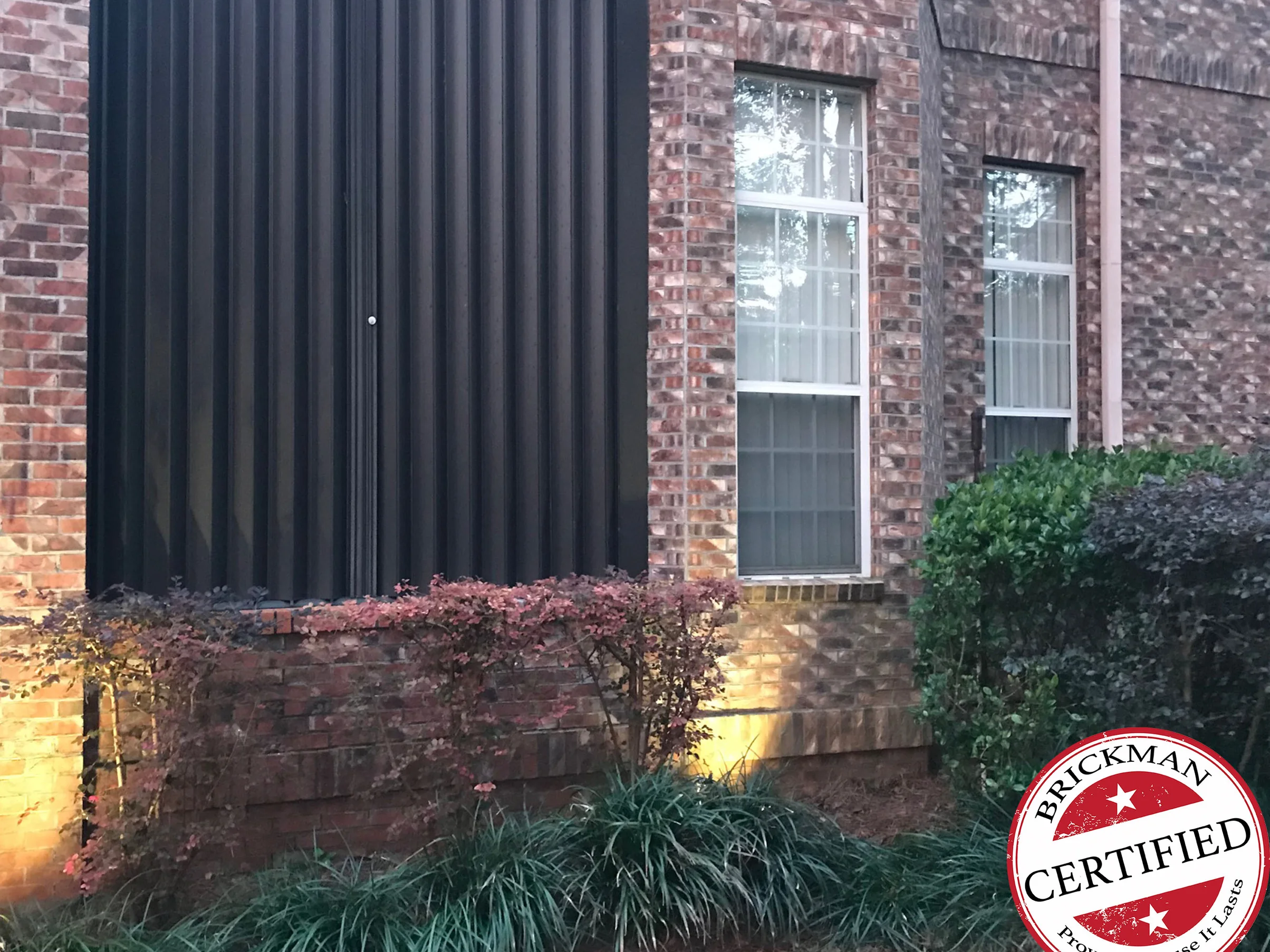 The same corner in 2021, the low wall and pier carrying the same brown and tan blend as the house above them, under a dark metal screen and behind low shrubs, with the certification stamp in the corner