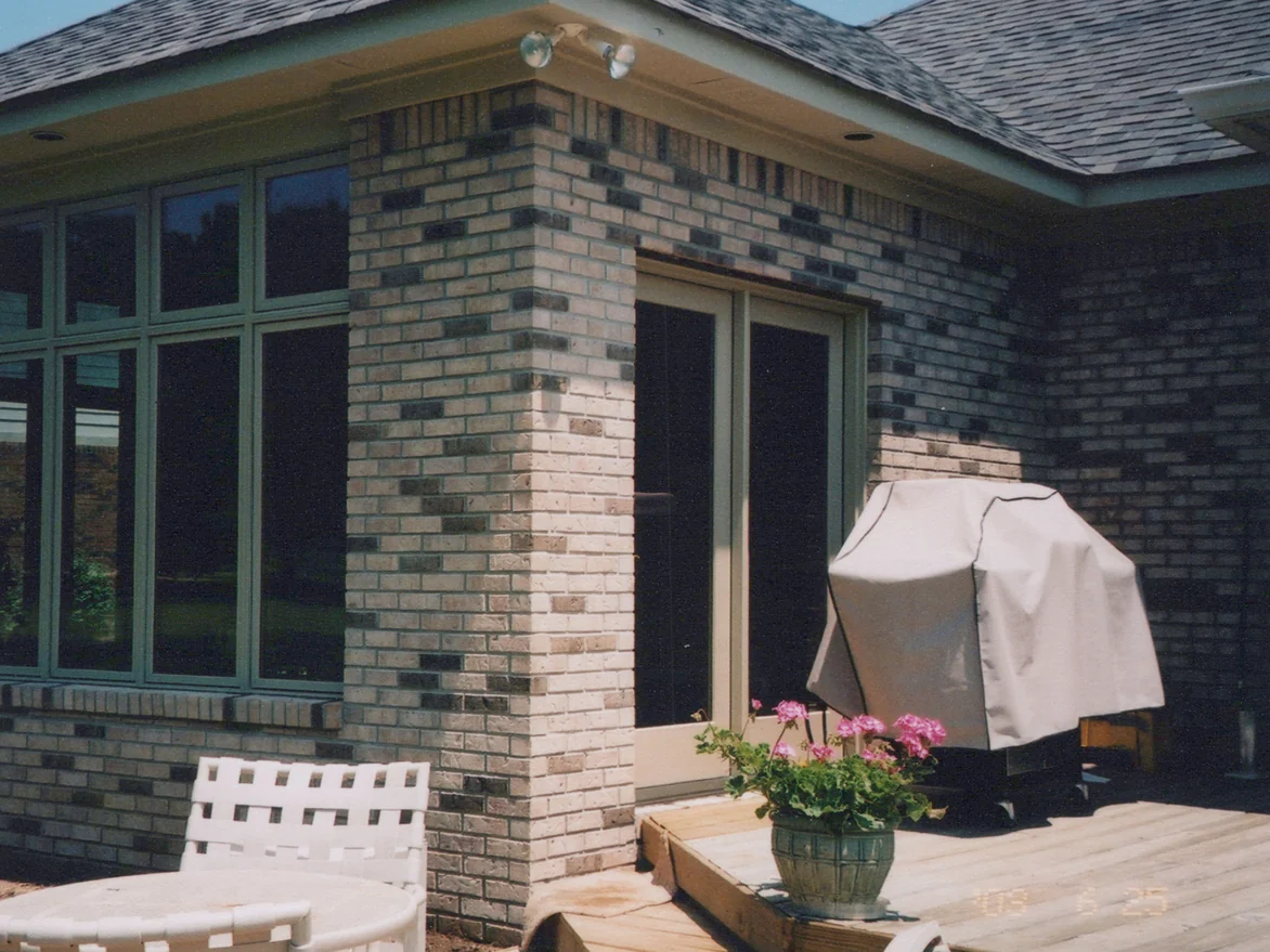 The corner of the addition in June 2003, its brick pier between a bank of windows and a glass door, with a covered grill and a potted geranium on the deck