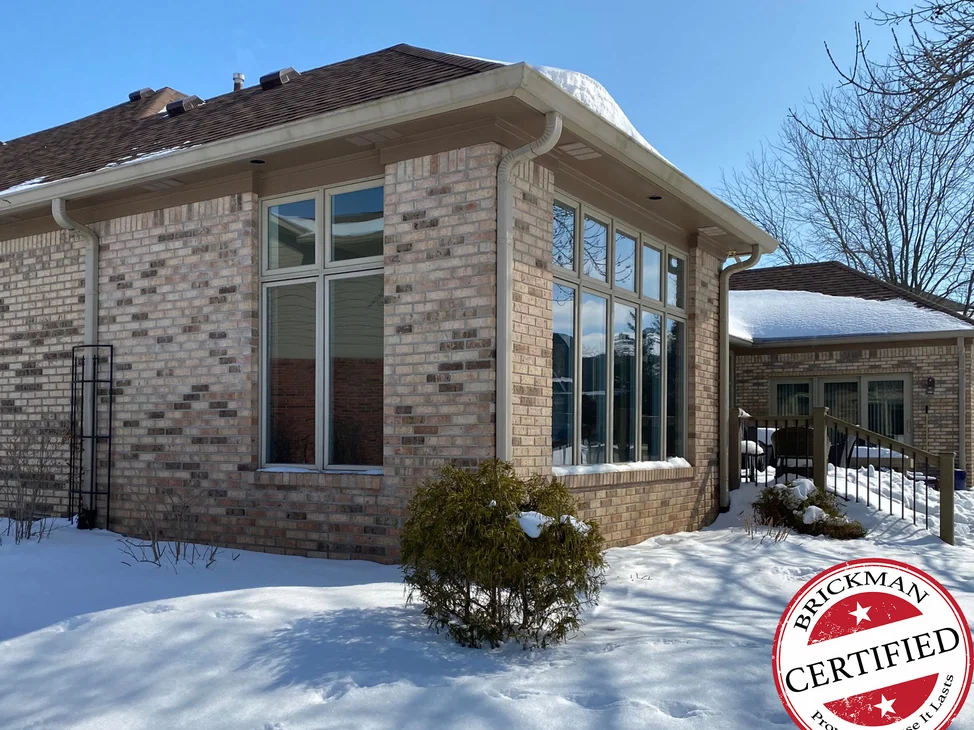 The corner of the addition in 2021 with snow on the ground, tan and brown brick running from the house wall on the left across the corner pier to the bank of windows, with the certification stamp in the corner