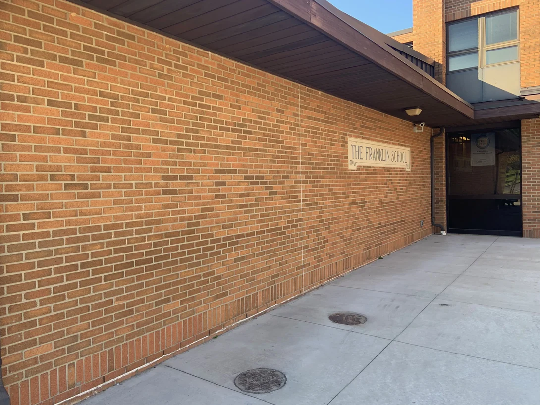 The entrance wall in 2019 under its deep timber soffit, a cast stone panel reading The Franklin School set in evenly blended brick