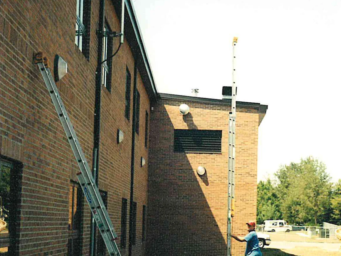A two story school wall in 2000 with an extension ladder and a pole standing against it, the brick a pale uneven tan