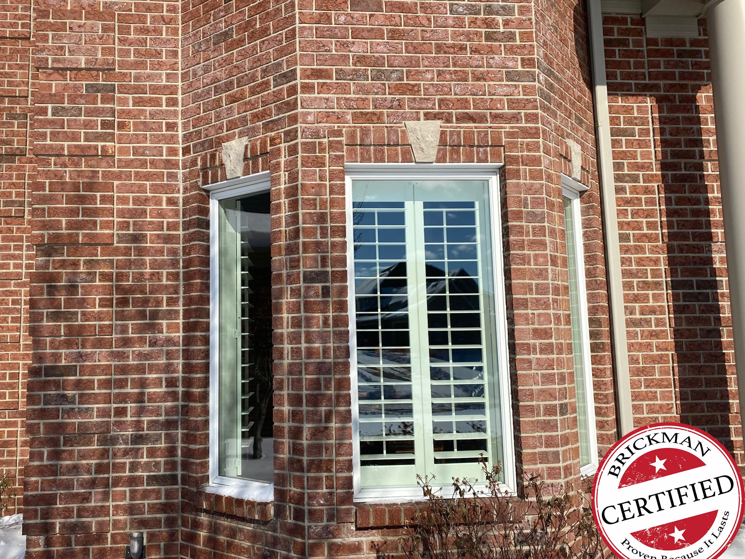 The brick bay window in winter 2021 with snow on the sills and shutters in the windows, red brown brick reading as one range under the stone keystones, with the certification stamp in the lower right corner