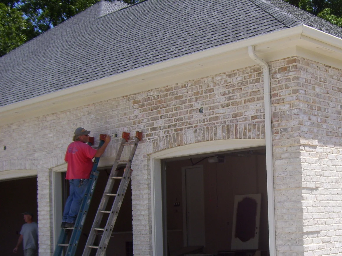 An applicator on a ladder in 2011, stain pots hooked to the rungs, working across the brick above the garage openings