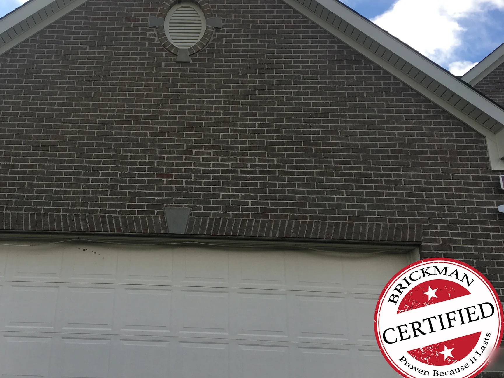 The garage gable in 2020, grey brown brick from the oval vent down to the soldier course and keystone above the white garage door, with the certification stamp in the lower right corner