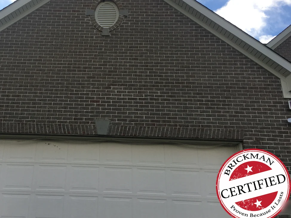The garage gable in 2020, grey brown brick from the oval vent down to the soldier course above the white garage door, with the certification stamp in the lower right corner