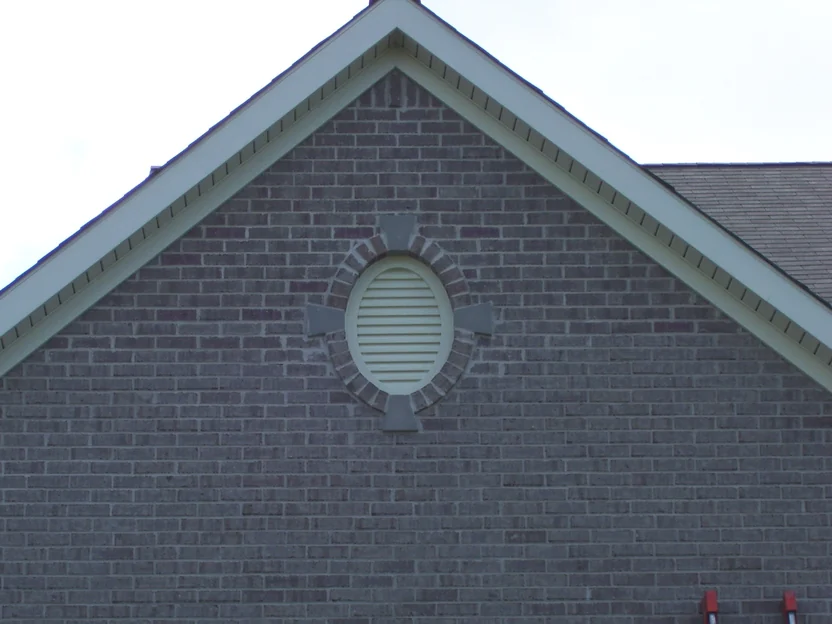 The garage gable in August 2007 under an overcast sky, grey brick with a red tinge on scattered units, an oval louvered vent set in a ring of brick under the peak and a ladder against the wall at right