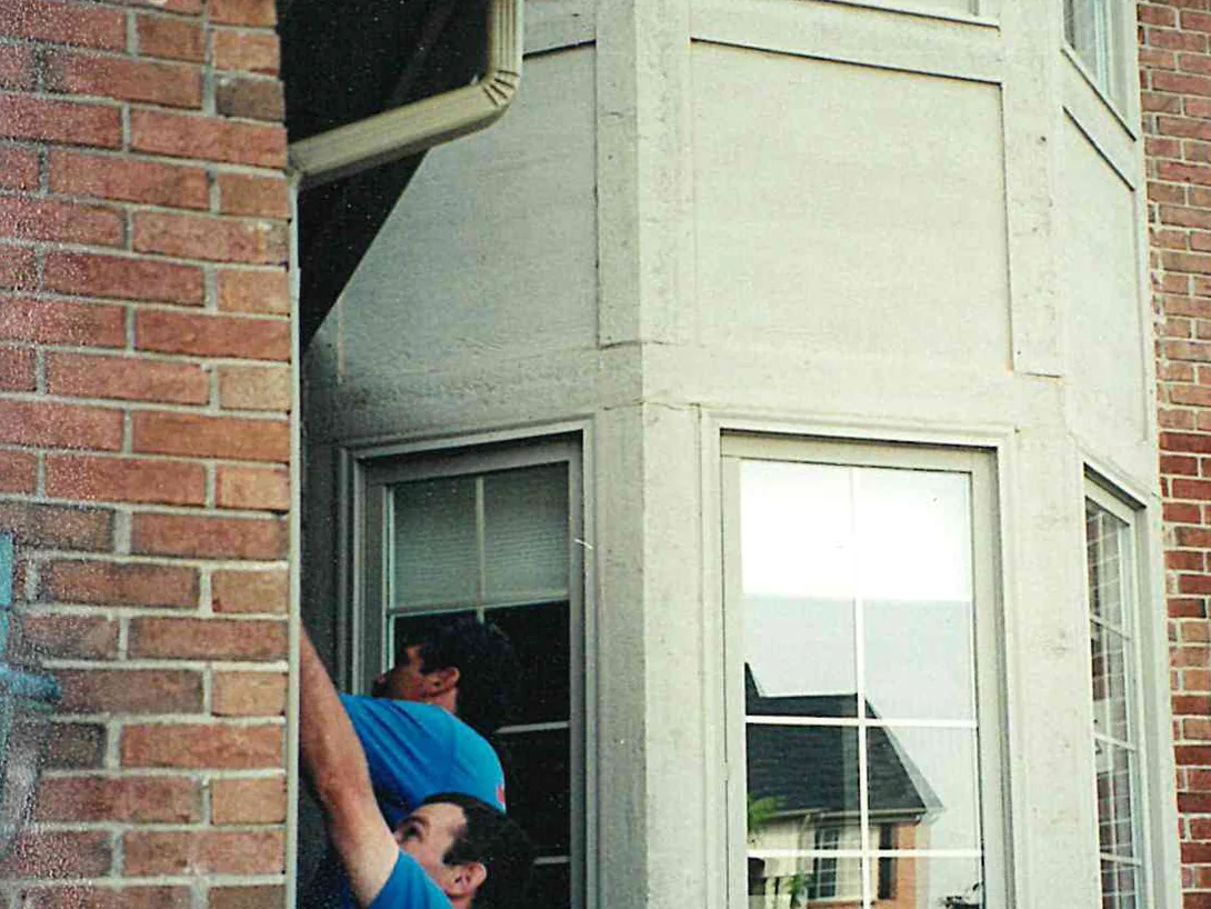 Two applicators working at the foot of a bay window, the brick pier beside them mottled red and tan