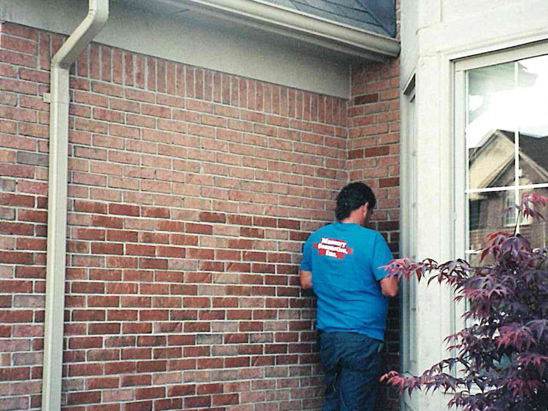 An applicator working along the brick of the house in 1999, patches of darker red showing across a lighter tan wall