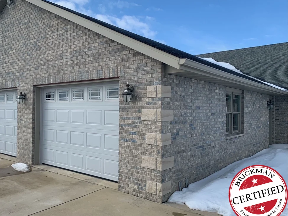 The garage corner of the Grand Mound house in 2021, grey and tan brick in one range across the gable and the return wall, with cut stone quoins and the certification stamp in the corner