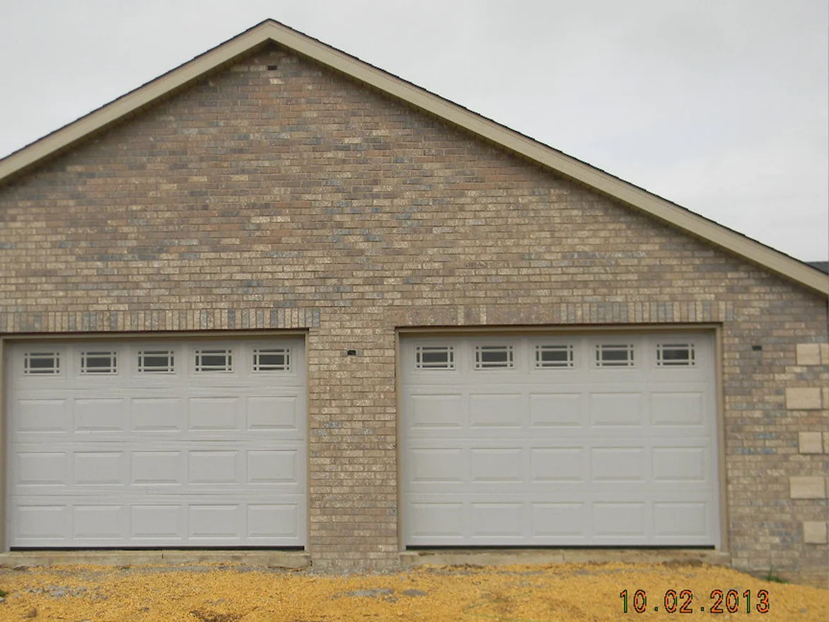 The garage gable of the house in October 2013 before staining, two white doors under a wall of tan brick blotched with darker units, bare dirt in front and a camera date in the corner