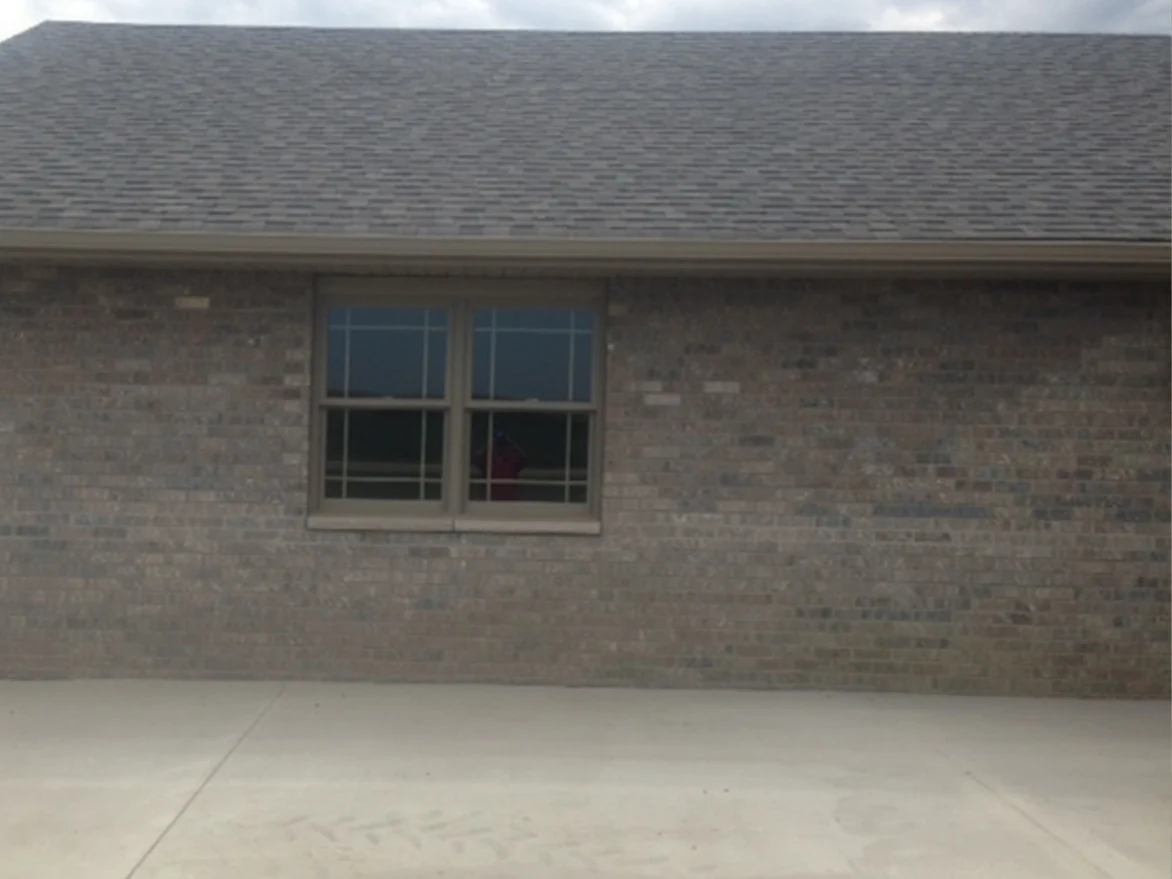 A side wall of the house before staining, one double-hung window in a wall of tan brick with a scatter of darker and lighter units, under a grey shingle roof