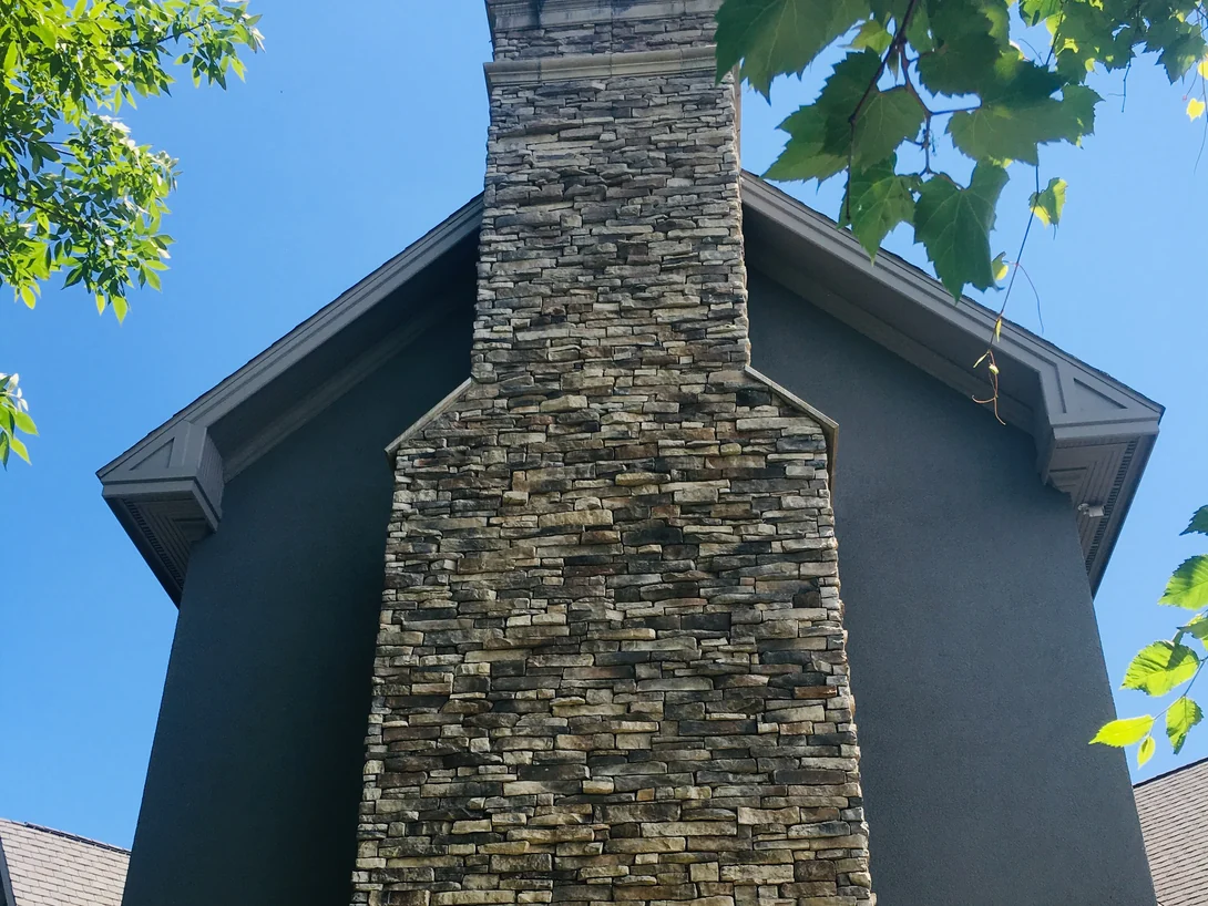The chimney from the ground in 2019, its corner returns holding the same tan and gray range as the face for the full height of the gable