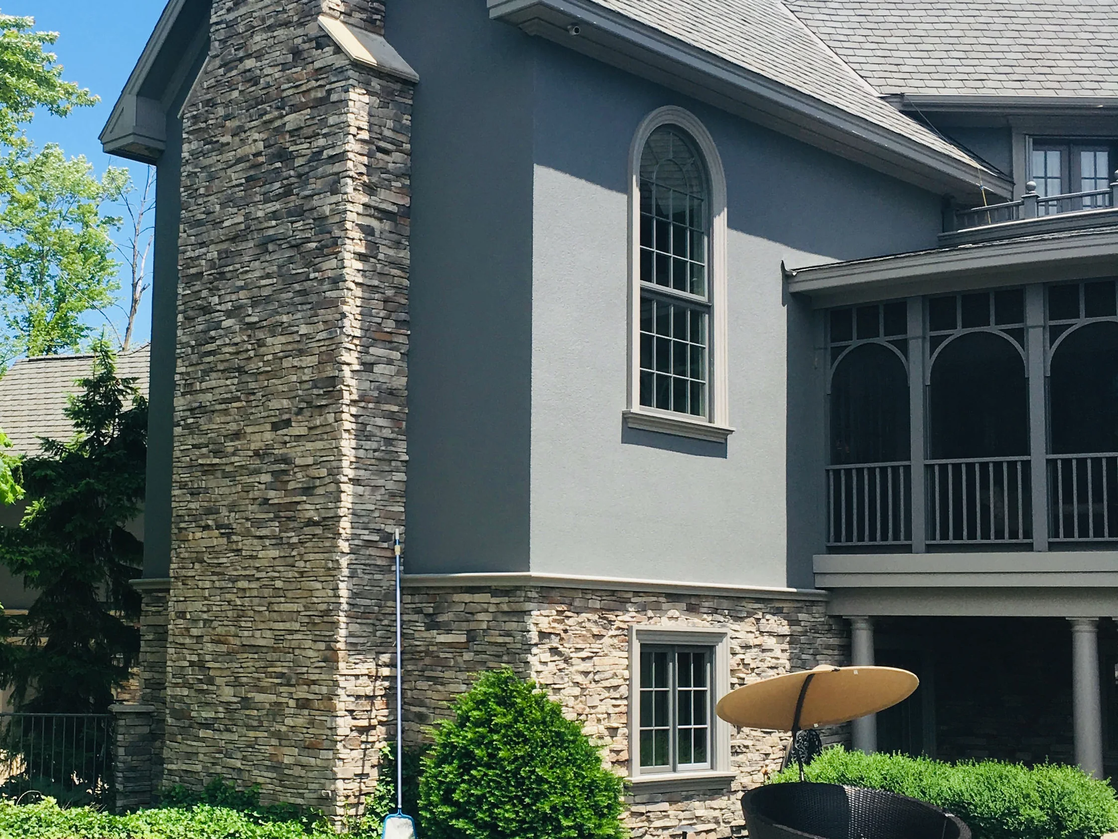 The gable end of the house in 2019 from the poolside, the stone chimney and the stone base course carrying one range of tan, gray and brown against gray stucco
