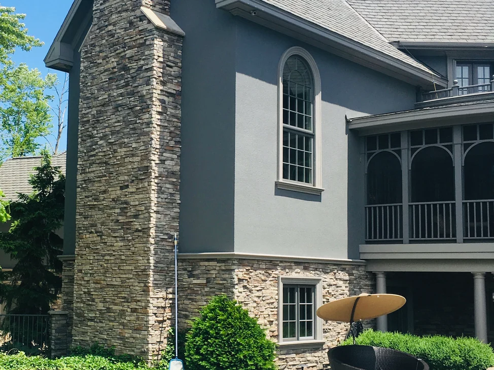 The gable end of the house in 2019 from the poolside, the stone chimney and the stone base course carrying one range of tan, gray and brown against gray stucco