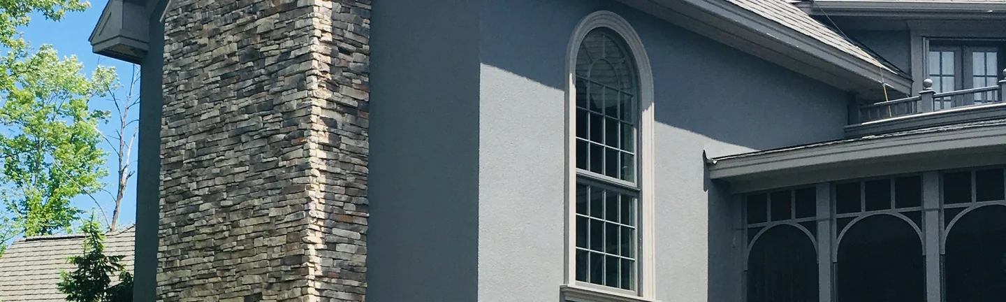 The stone chimney of a Grand Rapids house in 2019, running the full height of a gray stucco gable beside a tall arched window