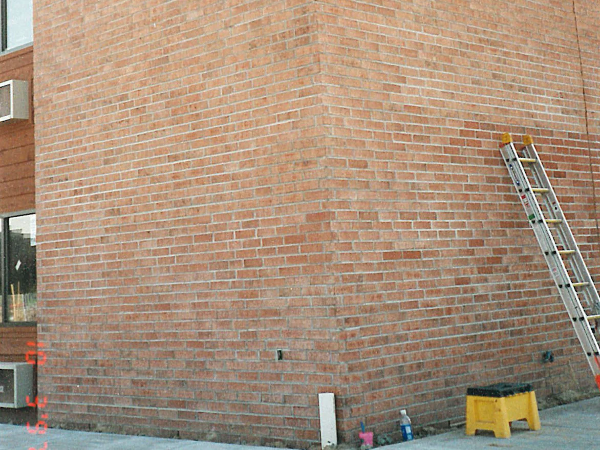 An outside corner of the hotel in 1997 with an extension ladder against the wall and a tub, a bottle and a step stool on the pavement below it