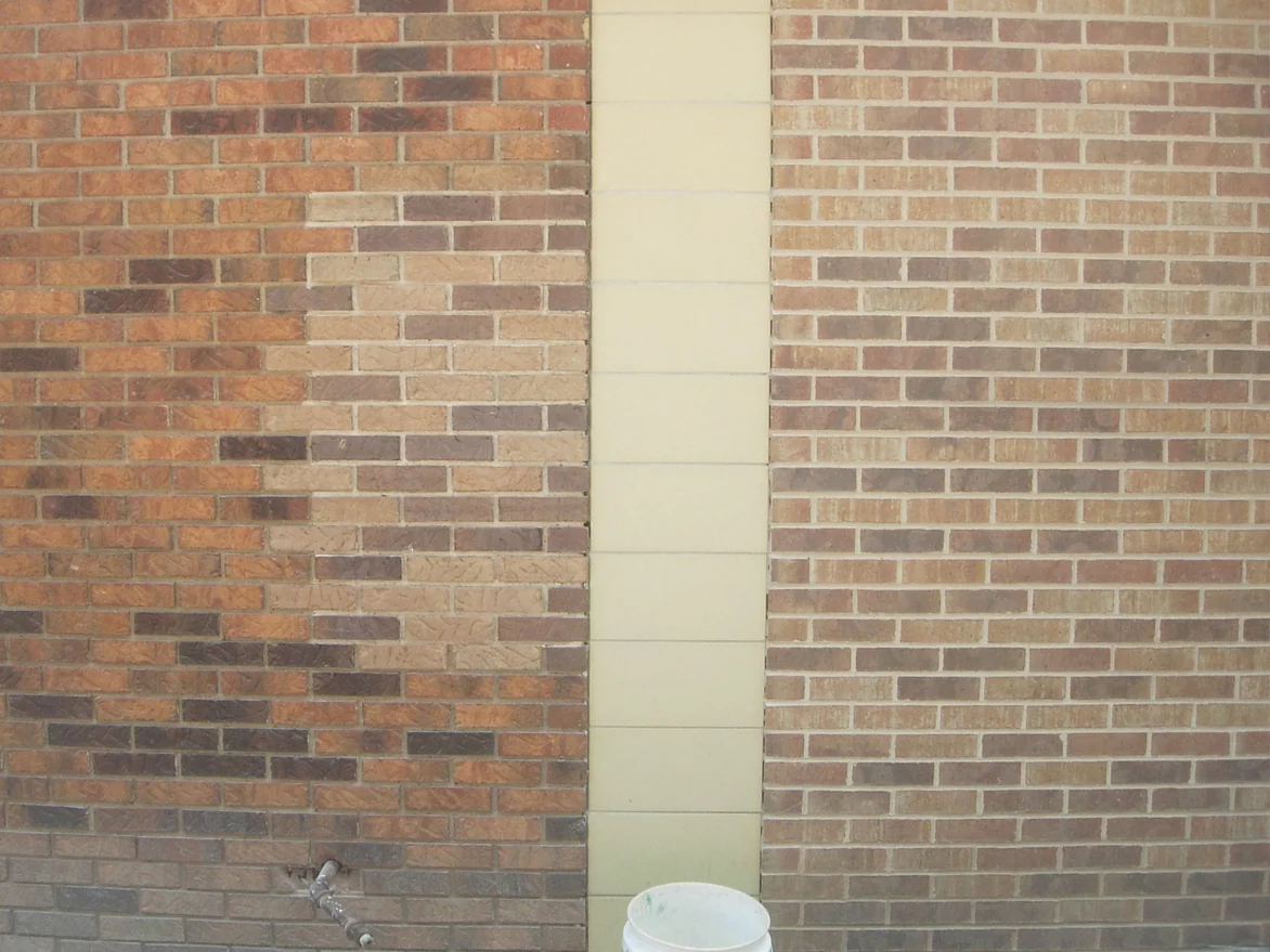 The wall mid-job in 2008, a patch of units on the textured brick already carried to a lighter range beside the untouched run, a cream block pier and the smooth tan brick to its right, a bucket at the foot