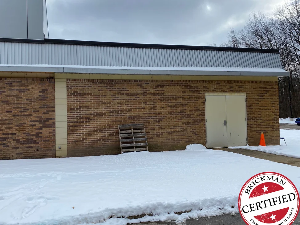 The side wall of the school wing in 2021, one range of tan and brown brick between cream block piers under a corrugated metal fascia, with the certification stamp in the corner