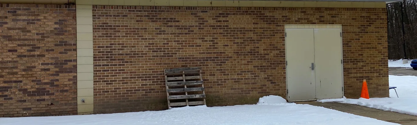 The side wall of the school wing in 2021, one range of tan and brown brick between cream block piers under a corrugated metal fascia