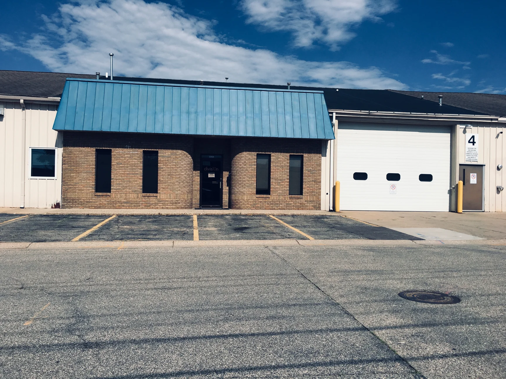 The whole office front from across the street in 2019, tan brick with two curved corners flanking a glass door under a blue standing seam mansard, a white overhead door on the bay next to it
