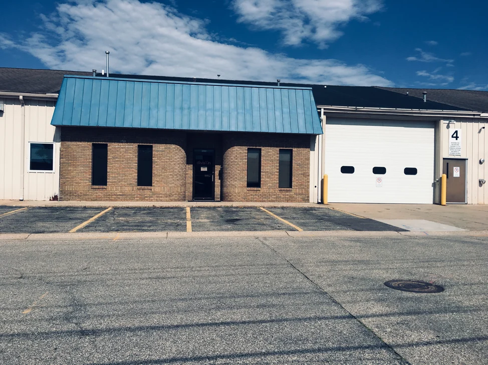 The office from across the street in 2019, tan brick with two curved corners flanking a glass door under a blue standing seam mansard roof