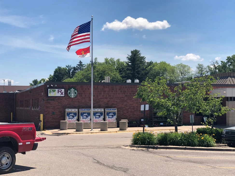 The front of the grocery store in 2019, one red brick wall running past a pharmacy sign and a coffee counter sign to a line of propane cages.