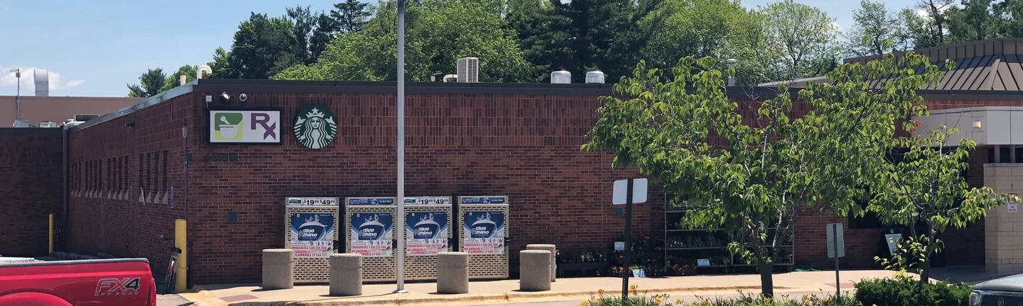 The front of a single-story grocery store in 2019, one red brick wall running the length of the frame behind a flagpole