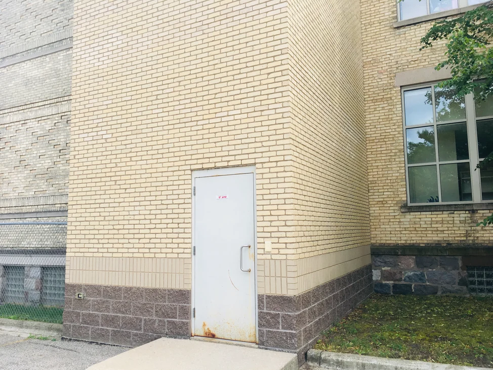 The foot of the elevator shaft in 2019, pale buff brick above a band of brown split face block, a grey steel door and a concrete ramp, with the older tan brick of the school on either side