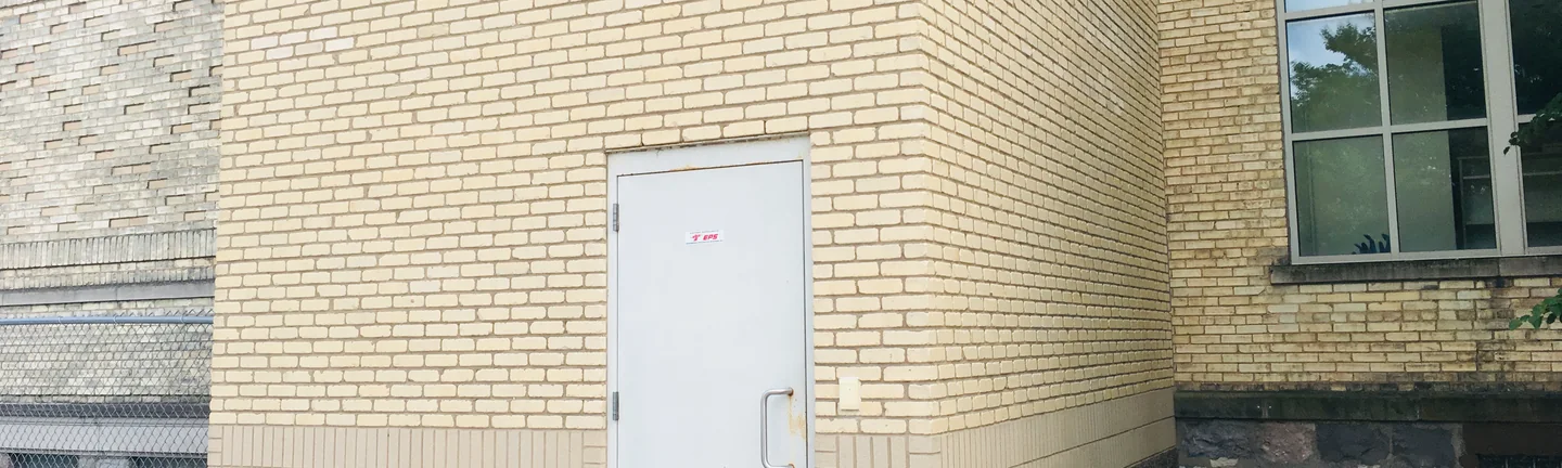 The foot of the elevator shaft in 2019, pale buff brick above a band of brown split face block, a grey steel door and a concrete ramp, with the older tan brick of the school on either side