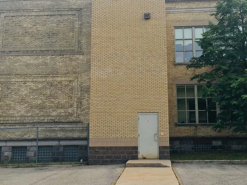 The elevator shaft in 2019 from the parking lot, its light brick running from the block base to the roofline between the older school walls, with a window bay and a tree at the right