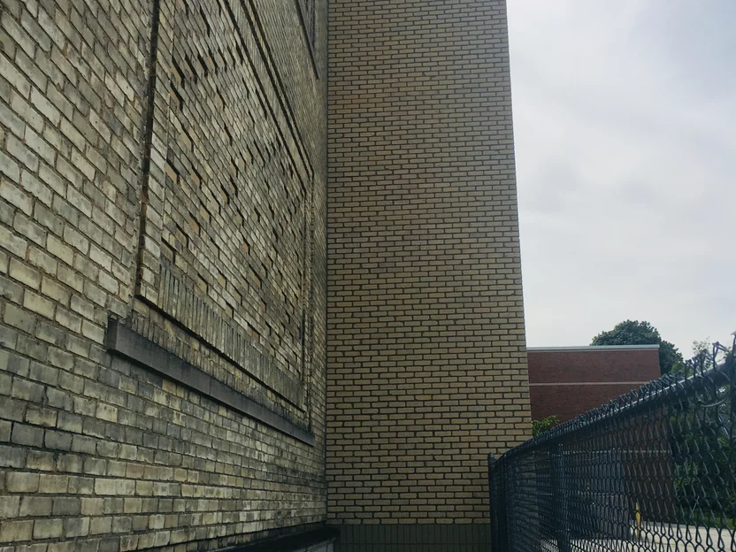 Looking along the side of the school in 2019, the older mottled tan brick at left and the light brick of the shaft standing forward of it, with a chain link fence and sky at the right