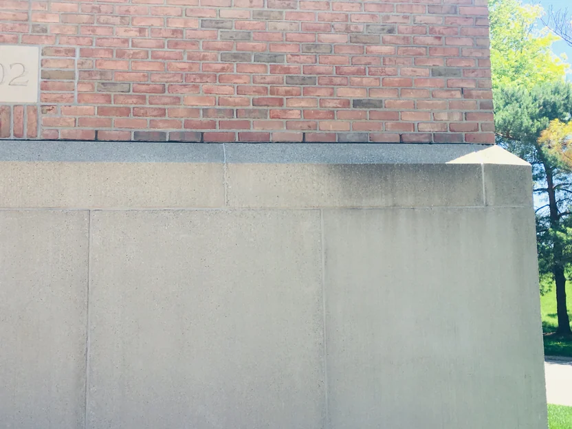 The bottom courses of the column in 2019 meeting the cast stone base, the edge of the date stone at the left and evergreens beyond the corner
