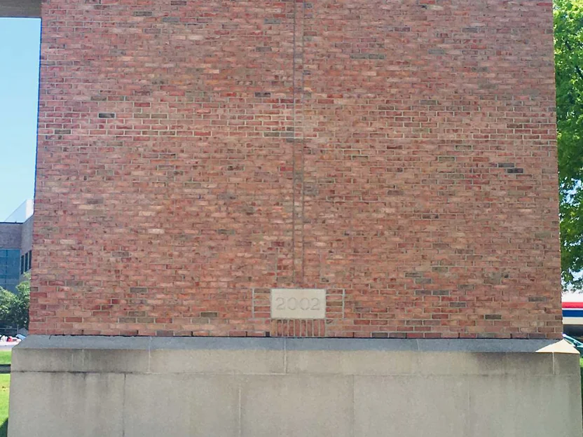 The full face of the brick column in 2019 under a soffit, salmon red brick running down to the 2002 date stone and the cast stone base, a tree at the right edge