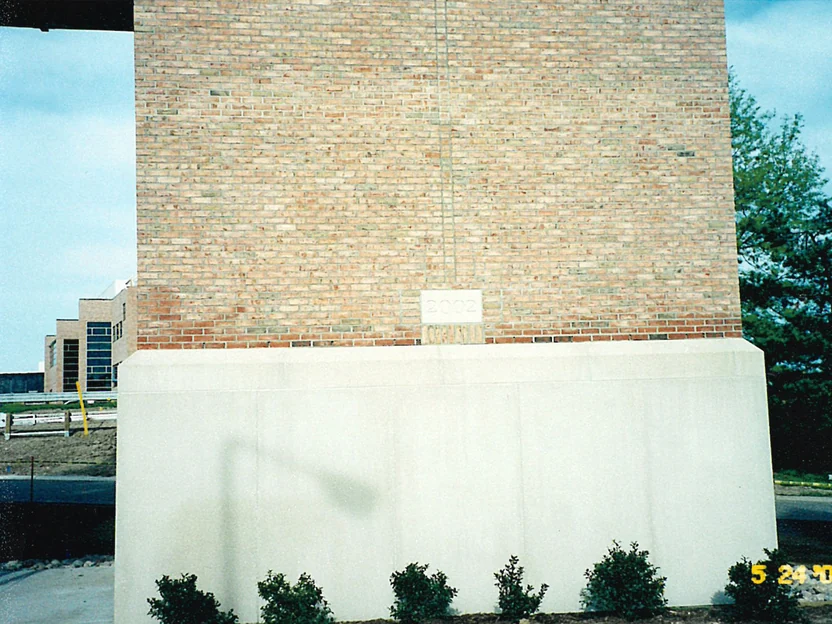 The brick column in May 2002, its face reading pale buff and tan above a band of redder brick at the 2002 date stone, on a fresh cast stone base with new shrubs in front and a camera date of 5/24/02 in the corner