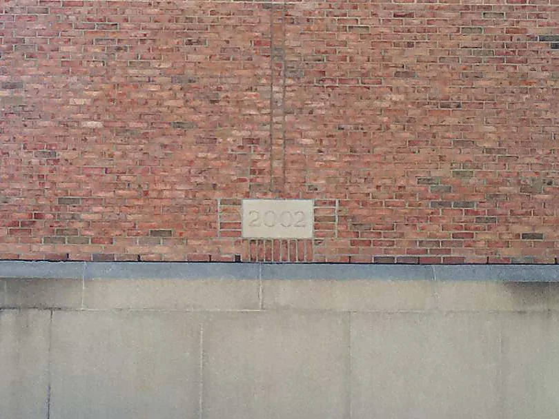 The 2002 date stone in 2019, set in salmon red brick just above the cast stone base, the courses around it holding one range
