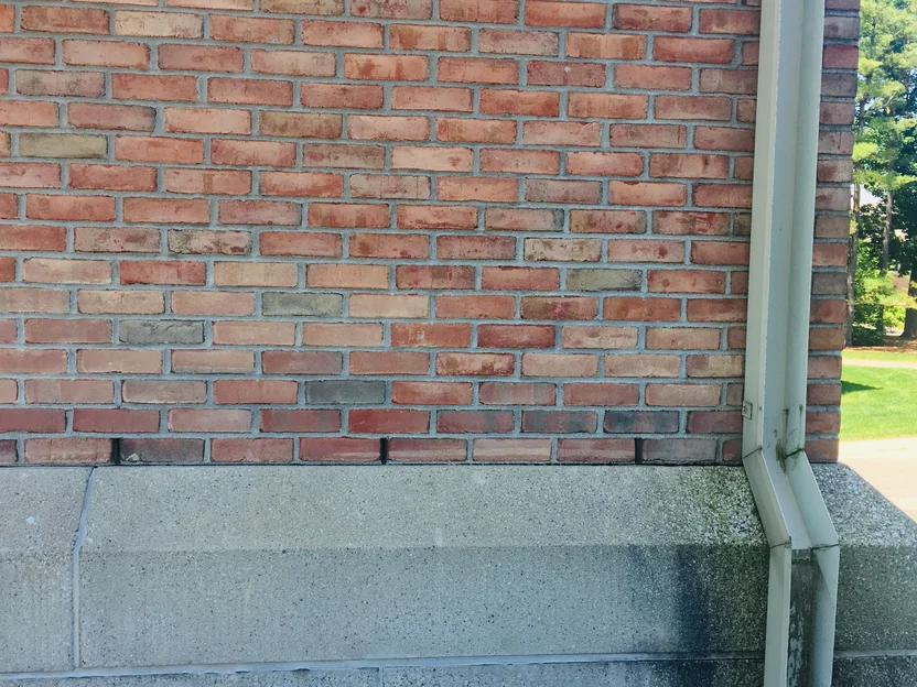 Brick at close range in 2019 beside a downspout, salmon, red and grey units in every course with grey mortar, over a cast stone base
