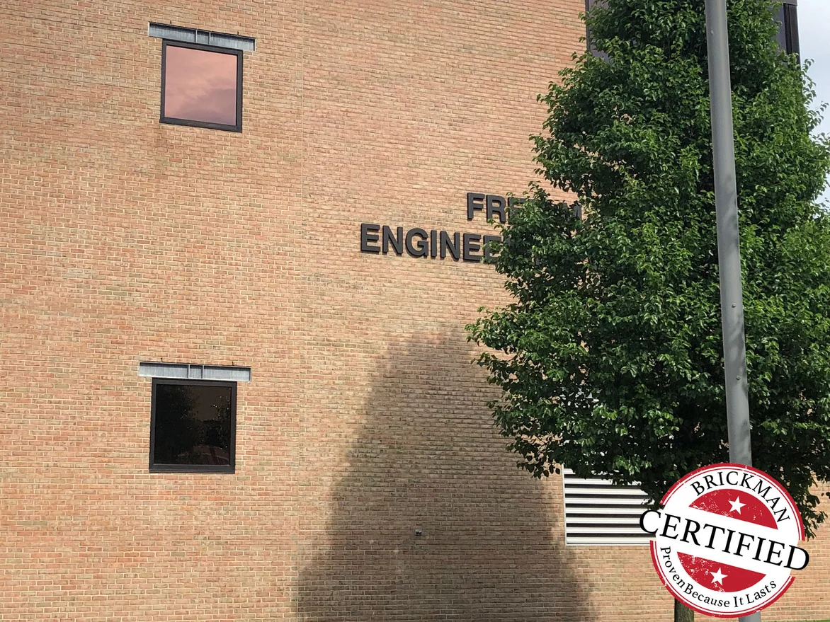 The same end wall in 2019 from the same side, the two stacked square windows and the louvre vent set in warm tan brick that holds one range across every course, the frame carrying the Brickman Certified stamp
