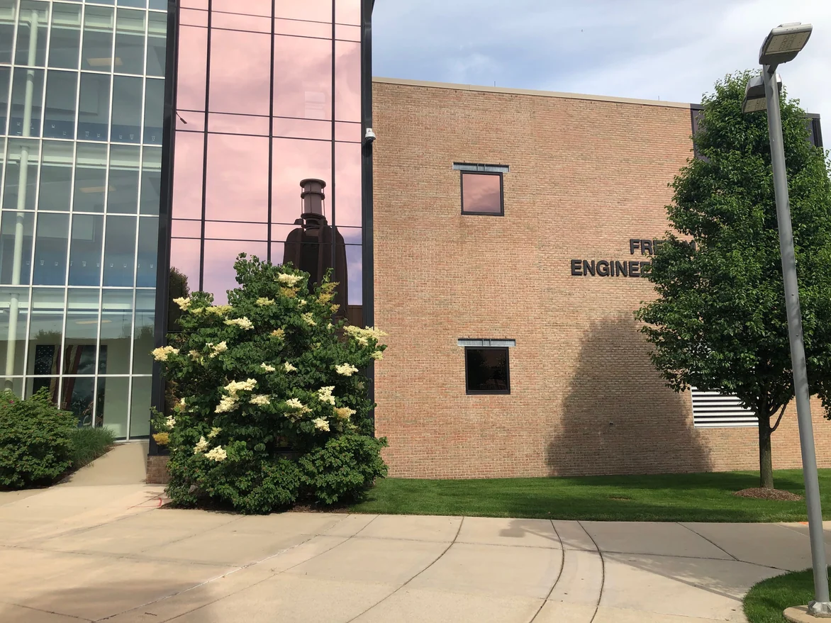 The engineering laboratories in 2019, a blank warm tan brick end wall meeting a full height glazed stair tower, with shrubs and a lamp standard at the base