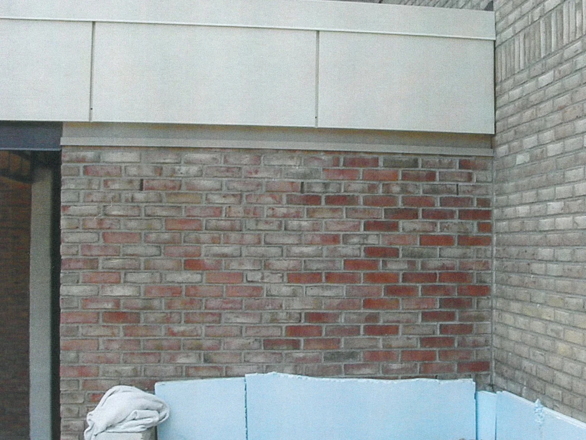 A red brick panel meeting the buff brick of the return wall at an inside corner in 2000, its units running from pale cream to dark red course by course, with foam board and sand at the base