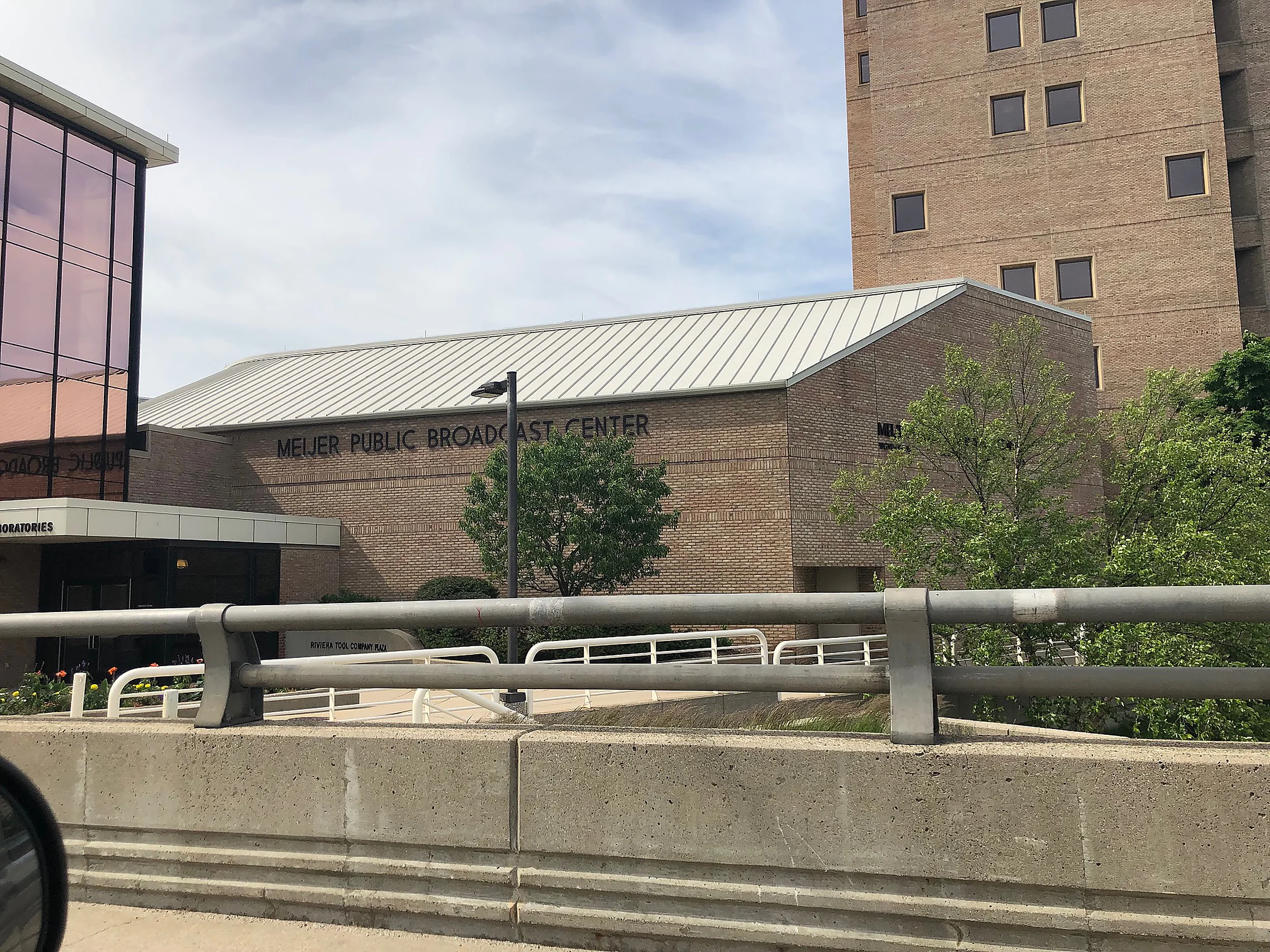 The Meijer Public Broadcast Center from the road in 2019, its lettered buff brick gable under a standing seam roof, with a taller brick block behind it and a glazed wing to the left