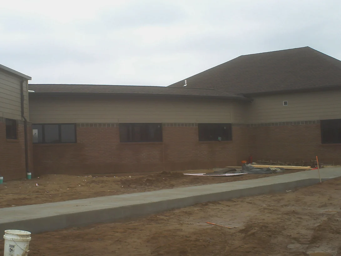 Two wings of the single storey building under construction around a courtyard of bare earth, a new kerb and walk poured and the masonry base course finished under lap siding