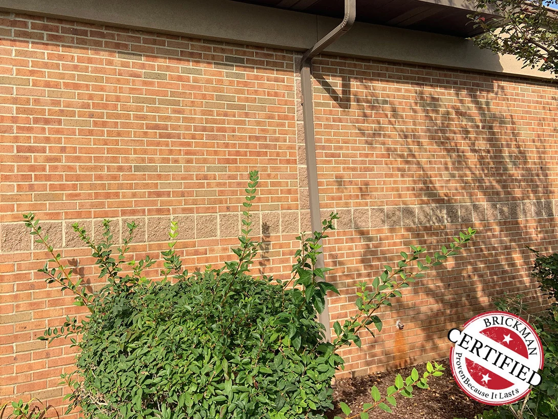 A wall under a deep eave in 2019 with a downspout running down it, the split faced block course crossing behind the shrubs and the brick above it even from end to end