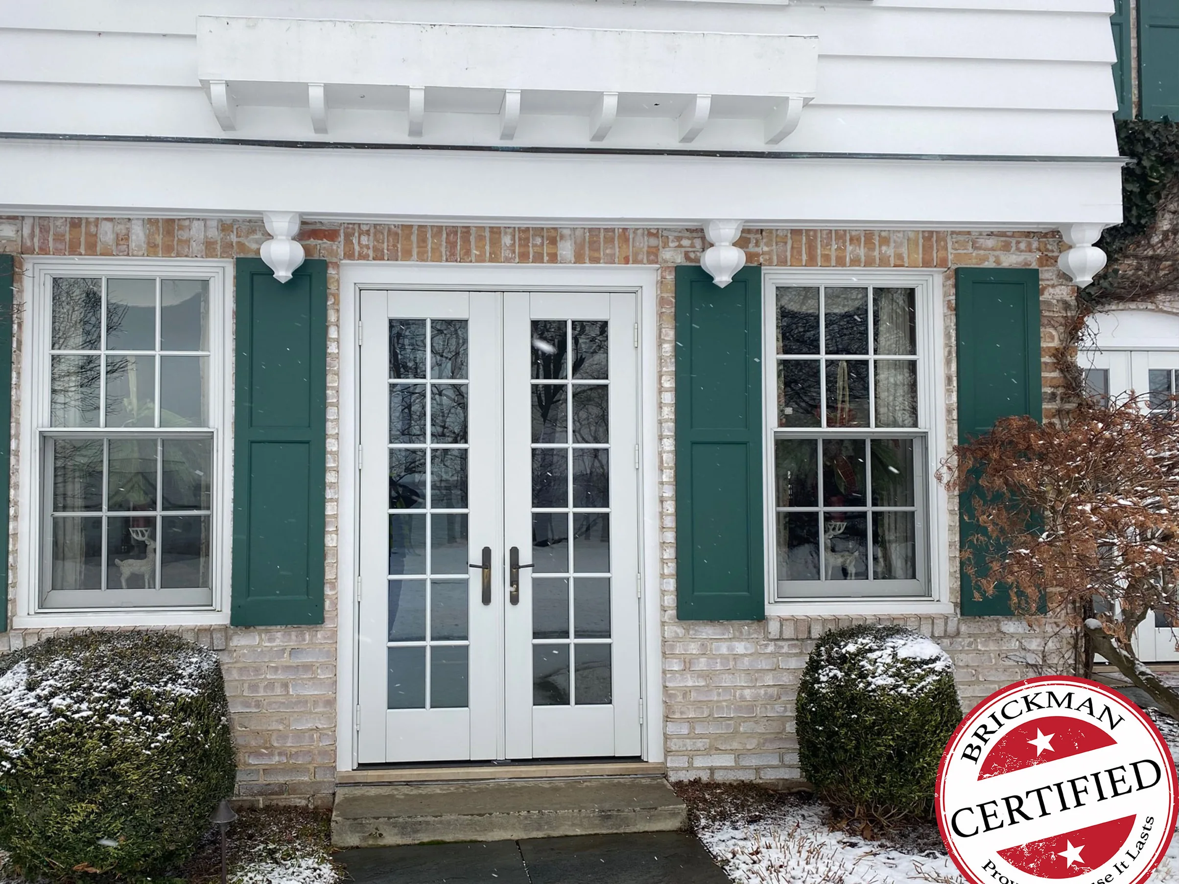 The French door entrance in 2021, white brick between green shutters under a white clapboard upper floor, snow on the boxwoods and the step, with the certification stamp in the corner